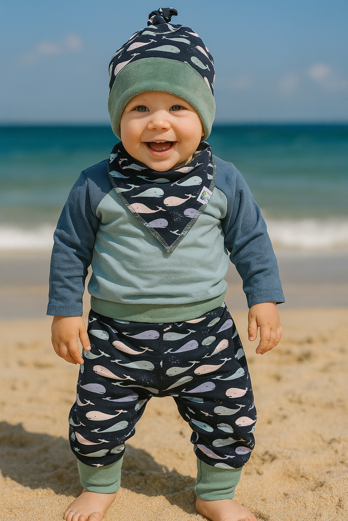 Whale Print Top Knot  hat. Child wearing a blue and green outfit with a Whale patterned hat and Bib on a beach.